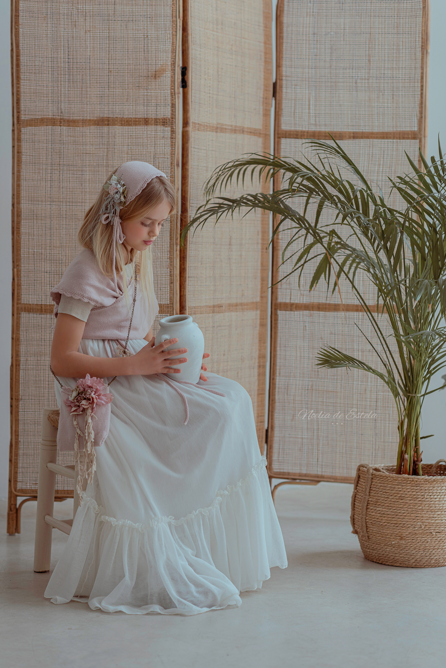 Niña sentada con vestido de Comunión y complementos rosas junto a un biombo